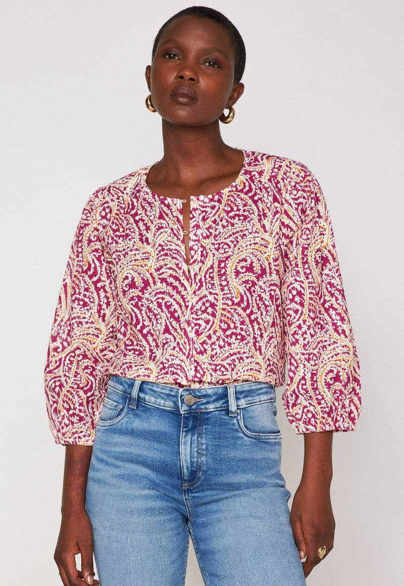Blusa a fiori rosa e bianca con maniche a tre quarti, scollatura rotonda e patta frontale. Abbinata a jeans blu a vita alta.
