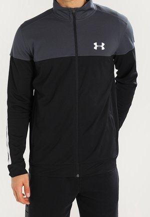 Mann, der eine schwarze und dunkelgraue Under Armour Jacke mit Reißverschluss und hohem Kragen trägt, steht vor einem schlichten Hintergrund.