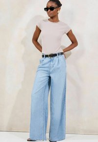 Camiseta ajustada de color beige claro, pantalones de mezclilla anchos de color azul claro, cinturón negro con hebilla dorada y gafas de sol oscuras. Tela suave y corte relajado.