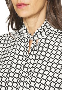 Blusa bianca con motivo geometrico nero, dotata di un colletto annodato e texture morbida. Dettagli in primo piano attorno all'area del collo.