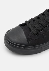 Schwarze Leinensneaker mit gummierter Zehenkappe, runden Schnürsenkeln und strukturiertem, schwarzem Gummiaußensohle, die auf der Kappe eine glatte Oberfläche aufweist.