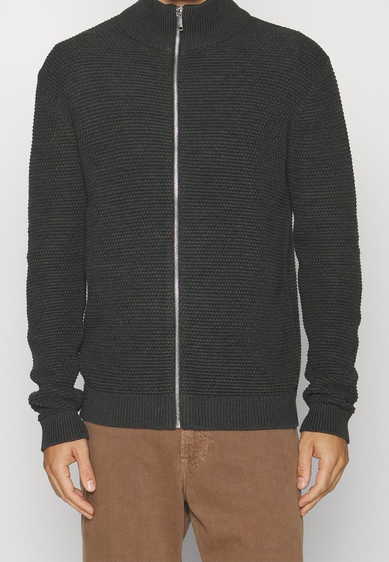Mörkgrå zip-up-tröja med hög krage, texturerad stickad mönster, ribbade cuffs och fåll, i kombination med ljusbruna byxor.