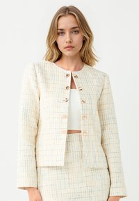 Vrouw in een crèmekleurige textuurjack met gouden knopen over een witte crop top en bijpassende rok tegen een effen achtergrond.