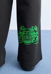 Sorte sweatpants med løs pasform, der har et skarpt grønt grafisk design af tegneserieagtige katte på den nederste venstre ben.