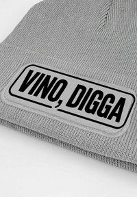 Gray knitted beanie featuring a rectangular patch with the text "VINO, DIGGA" in bold black lettering on a white background. Textured fabric.