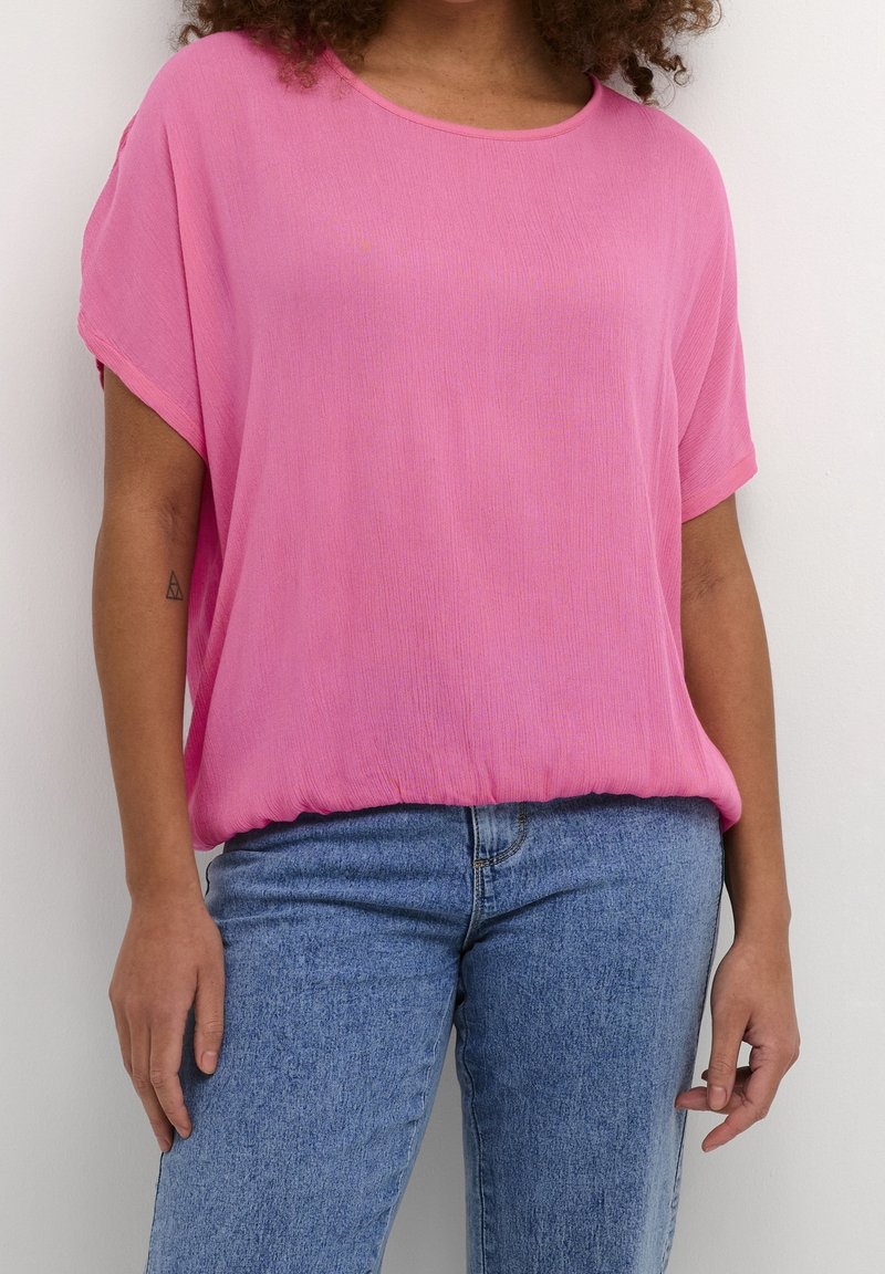 Blusa rosa, holgada, con cuello redondo y mangas cortas, hecha de tela ligera. Combinada con jeans de pierna recta en azul claro.
