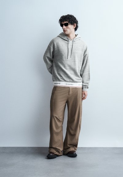 Uomo con capelli ricci che indossa occhiali da sole neri, felpa grigia con cappuccio, pantaloni marroni a gamba larga e scarpe nere in piedi contro una parete azzurro pallido.