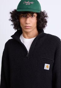 Črna pletena pulover s zip ovratnikom in sprednjim žepom. Ima izstopajoč oranžni logotip. Model nosi zeleno kapo.