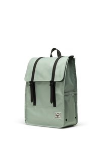 Sac à dos vert menthe avec une fermeture à rabat, des sangles noires et une poche avant. Fabriqué en tissu durable, présentant une forme rectangulaire et un patch logo.