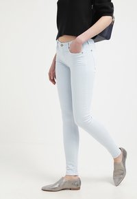 Jeans ajustados de color azul claro con una textura suave, botón plateado y dos bolsillos frontales, combinados con zapatos grises sin cordones. Fondo blanco.