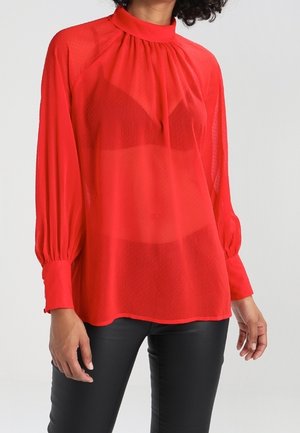 Vrouw die een doorschijnende rode blouse met lange mouwen en een hoge hals draagt, gecombineerd met een zwarte broek, tegen een effen lichte achtergrond.