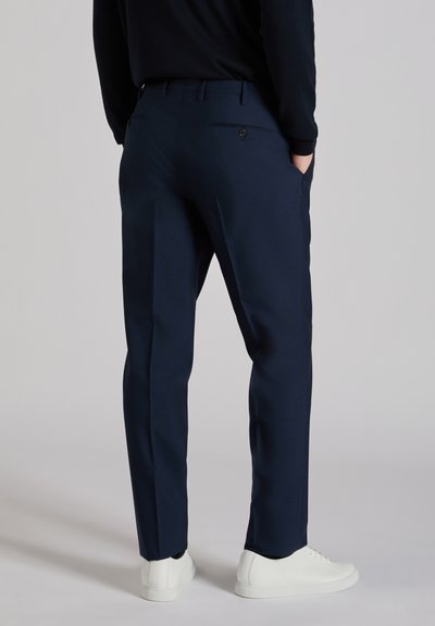 Pantalon tailleur bleu marine à la texture lisse, avec une poche arrière à bouton et une silhouette élégante, porté avec des baskets blanches.