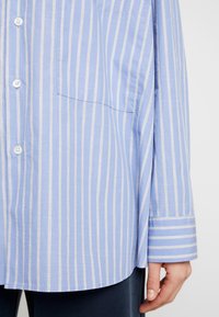 Světle modrá košile na knoflíky vyrobená z měkké látky, s vertikálními bílými pruhy, přední kapsou a dlouhými rukávy s manžetami a pruhovaným detailem.