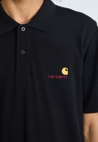 Polo shirt noir en coton, avec un col classique, deux boutons et un logo Carhartt brodé en jaune et rose sur la poitrine gauche.