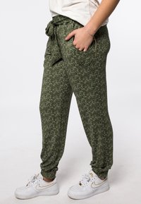 Pantaloni sportivi verdi a fantasia con vita elastica e lacci, dotati di tasche laterali e polsini a coste, abbinati a scarpe bianche.