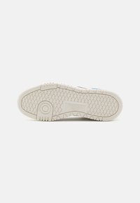 Podeszwa sneakerów Reebok w białej gumie z teksturowanym wzorem trakcyjnym, z okrągłym detalem w pobliżu pięty i wytłoczonym logo na środku.