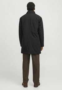 Cămașă neagră cu guler înalt și mâneci lungi, având un croi drept. Materialul pare neted și ușor. Asortată cu pantaloni marron.