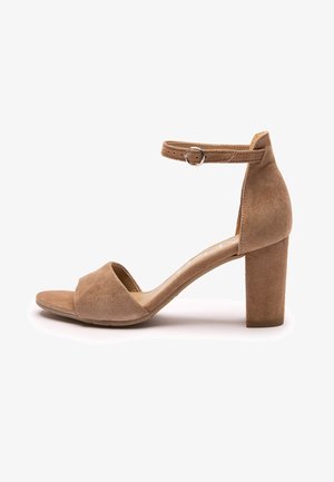 Brązowy sandał na obcasie zamszowy z odkrytymi palcami i regulowanym paskiem wokół kostki z srebrną klamrą, pokazany z boku na białym tle.