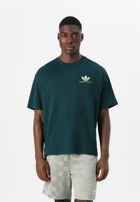 Donkergroen katoenen t-shirt met korte mouwen, voorzien van een gouden Adidas-logo en de tekst "BASKETBALL". Gecombineerd met lichtgrijze geraffineerde shorts.