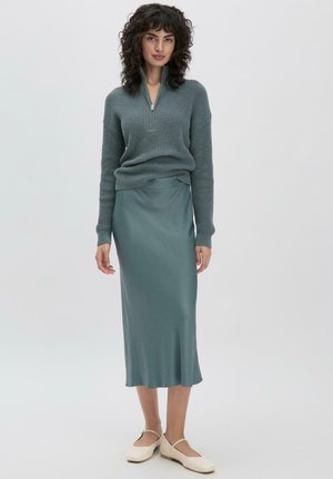 Türkiser Strickpullover mit einem Viertel-Reißverschluss und geripptem Muster, kombiniert mit einem satinähnlichen türkisfarbenen Midi-Rock. Das Model trägt cremefarbene Slipper.