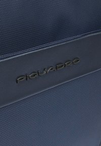 Primo piano di tessuto blu strutturato e sezione in pelle liscia con logo Piquadro nero in rilievo.