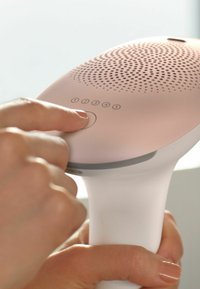 Dispositif portable avec une poignée blanche lisse et un dessus rose clair. Il comporte cinq boutons de contrôle, une zone de haut-parleur texturée et une forme arrondie.