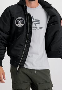 Musta bomber-takki hupulla, jossa Apollo 11 -merkki. Päällä vaaleanharmaa T-paita ja oliivinvihreät cargo-housut.