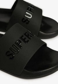 Zwarte slippers van textuur rubber, met een brede band die het woord "SUPER" in glanzende letters is gedrukt.