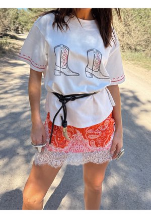 Wit T-shirt met cowboylaarzenprint, rode borduursels op de mouwen, gecombineerd met een oranje paisley rok met witte kantafwerking, zwarte riem.