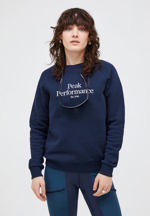Marineblauwe sweatshirt met het "Peak Performance" logo in het wit, met ribbels bij de mouwen en onderkant. Draagt over donkere, strakke broek met zakdetails.