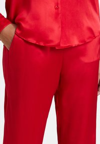 Ensemble de pyjama en satin rouge comprenant une chemise à boutons et un pantalon assorti ; texture lisse, coupe décontractée et poches latérales.
