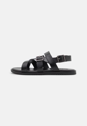 Black flat sandal with three leather straps and two silver buckles viewed from the side on a white background.