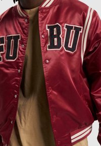 Veste bomber en satin rouge avec poignets et col côtelés rayés blancs, brodée des lettres "FU" et "BU" en noir sur la poitrine.