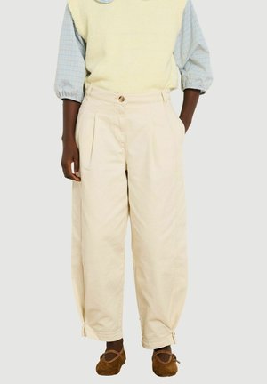Vrouw met beige wijde broek met zijzakken, een lichtgele gilet over een blauw geruit hemd en bruine platte schoenen.