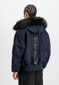 Marine pufferjack met een nep-bont capuchon. Heeft een glanzende textuur, elastische manchetten, zijzakken en gedurfde branding langs de achterkant.
