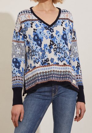 Kobieta ubrana w niebieski sweter z wzorem kwiatowym, z dekoltem w serek oraz granatowymi mankietami i wykończeniem u dołu, zestawiona z niebieskimi dżinsami.