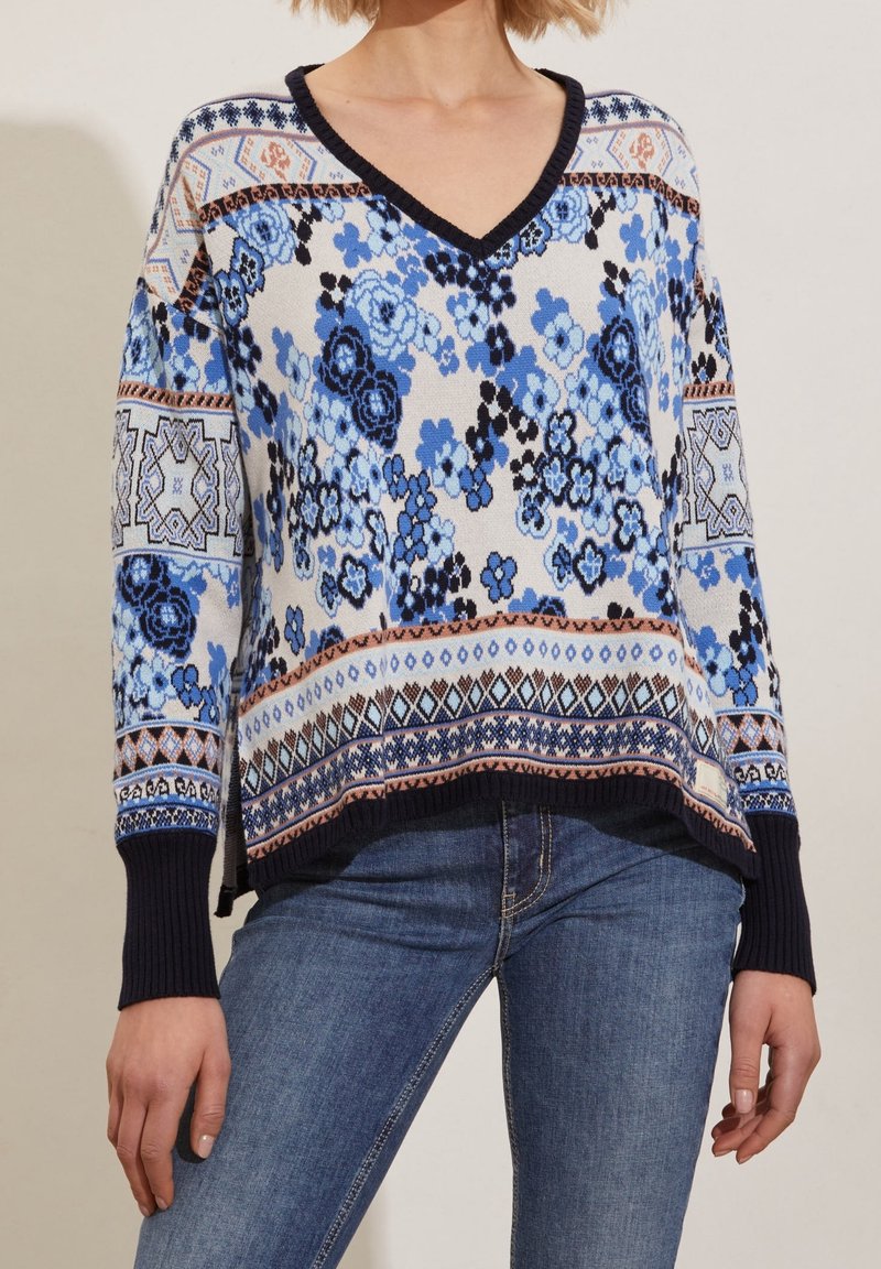 Femme portant un pull à col en V bleu à motif floral avec des poignets et un ourlet bleu marine, associé à un jean bleu.