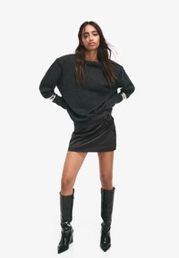 Nadrozmerný tmavosivý sveter so širokým výstrihom, kombinovaný s elegantnou čiernou mini sukňou s ružovými akcentmi. Modelka má na sebe vysoké čierne čižmy na opätku.