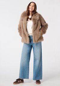 Veste en fausse fourrure marron avec des revers larges, associée à un jean large bleu clair et des baskets marron avec des rayures roses. Texture douce, coupe décontractée.