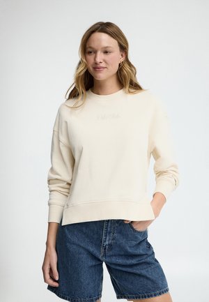 Vrouw met golvend blond haar, gekleed in een crèmekleurige trui met ronde hals en blauwe spijkershorts, staand met één hand in de zak tegen een effen achtergrond.