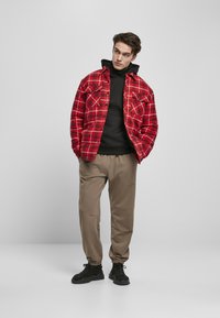 Rød rutete jakke over en svart hettegenser, beige tapered bukser og svarte sneakers. Jakken har knappefront og sidelommer.