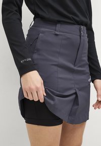 Grijze skort met voorplooien en zijzakken, gemaakt van een gladde stof. Draag het over zwarte fietsbroeken, met knoopsluiting en riemlussen.