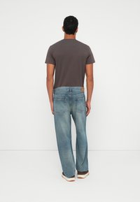 Bărbat îmbrăcat cu un tricou gri închis, blugi deschiși la culoare și încălțări maro, stând cu fața spre un zid alb, cu mâinile relaxate de-a lungul corpului.