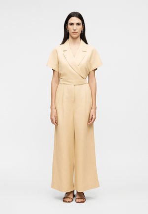 Γυναίκα με μακριά σκούρα μαλλιά, ντυμένη με μπεζ τοπ τύπου wrap με κοντά μανίκια και παντελόνι με φαρδιά μπατζάκια, που στέκεται ξυπόλυτη μπροστά σε απλό φόντο.