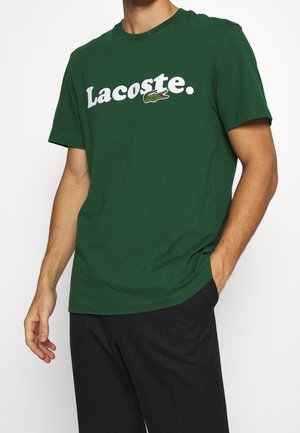 Mann trägt ein dunkelgrünes kurzärmeliges T-Shirt mit weißem "Lacoste"-Schriftzug und kleinem Krokodil-Logo auf der Brust, kombiniert mit schwarzen Hosen.