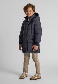 Polstret mørkegrå jakke med hætte, der har lynlåslommer og trykknaplukning, båret over beige bukser og sneakers.