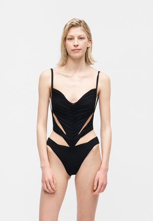 Moteris dėvi juodą ryškią vientisą maudymosi kostiumėlį su iškirptėmis šonuose ir plonomis petnešėlėmis, stovinti priešais šviesų paprastą foną.