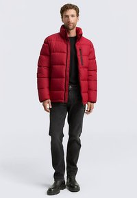 Chaqueta acolchada roja con cuello alto, bolsillo en el pecho con cremallera y detalles en negro. Se lleva sobre una camiseta negra y vaqueros oscuros, combinada con botas negras.