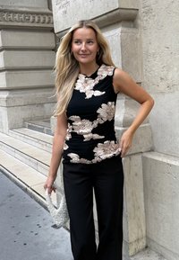 Fekete ujj nélküli felső, ráncolt mintával, amely nagy virágmintákkal van díszítve krémszínű és bézs árnyalatokban. Fekete, bő szárú nadrággal és fehér táskával párosítva.