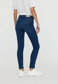 Jeans en denim bleu skinny avec cinq poches, taille mi-haute, et une étiquette en cuir à l'arrière de la ceinture. Portés avec des baskets blanches à plateforme.